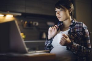 Pregnant woman thoughtfully looks at a computer while holding a white coffee cup