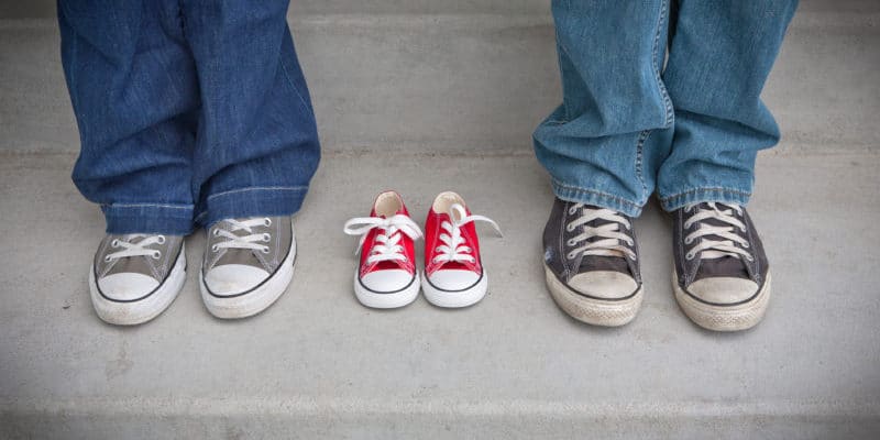 Two adult grey sneakers frame a small red pair of empty sneakers.
