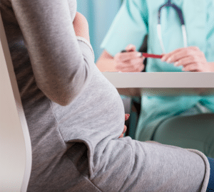 pregnant woman in a grey sweater dress sits across the table from a doctor