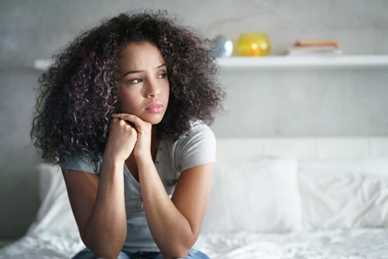 A woman with dark curly hair contemplatively looks away while sitting at the foot of her bed