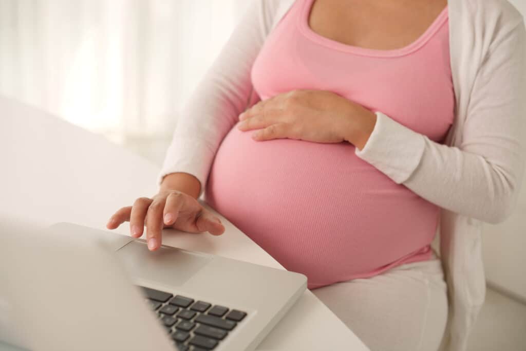 Pregnant woman in a pink tank top rests her hand over her belly while working on a laptop