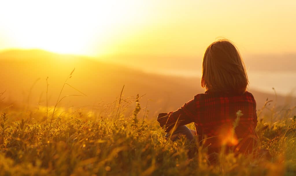 Woman wearing a plaid shirt sits in a field looking toward the sunset